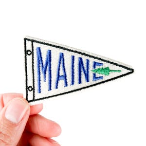 May include: A white pennant patch with the word "MAINE" embroidered in blue, and a green tree graphic. The patch has a black border and is held by a hand against a white background. The patch is triangular in shape.