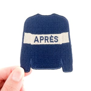 Pode incluir: Um patch bordado azul marinho em forma de camisola. O patch apresenta uma faixa horizontal branca com a palavra "APRÈS" em letras azuis marinho. O patch é segurado por uma mão contra um fundo branco.
