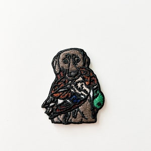 May include: Embroidered patch featuring a brown dog holding a colorful duck in its paws. The dog is wearing a green collar.