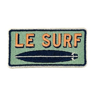Puede incluir: Un parche rectangular bordado con fondo verde claro y un gráfico de tabla de surf azul oscuro centrado bajo el texto naranja LE SURF. El parche tiene un borde azul oscuro y un acabado de borde cosido en color claro.