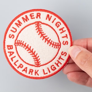 Op de afbeelding: Geborduurde patch met een honkbalontwerp. De patch heeft rode stiksels op een crèmekleurige achtergrond, met de woorden "SUMMER NIGHTS BALLPARK LIGHTS" in rood rond de honkbal.