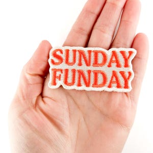 Puede incluir: Un parche rectangular con las palabras "SUNDAY FUNDAY" bordadas en hilo naranja brillante sobre un fondo de tela blanca. El parche se sostiene en la mano de una persona sobre un fondo blanco.