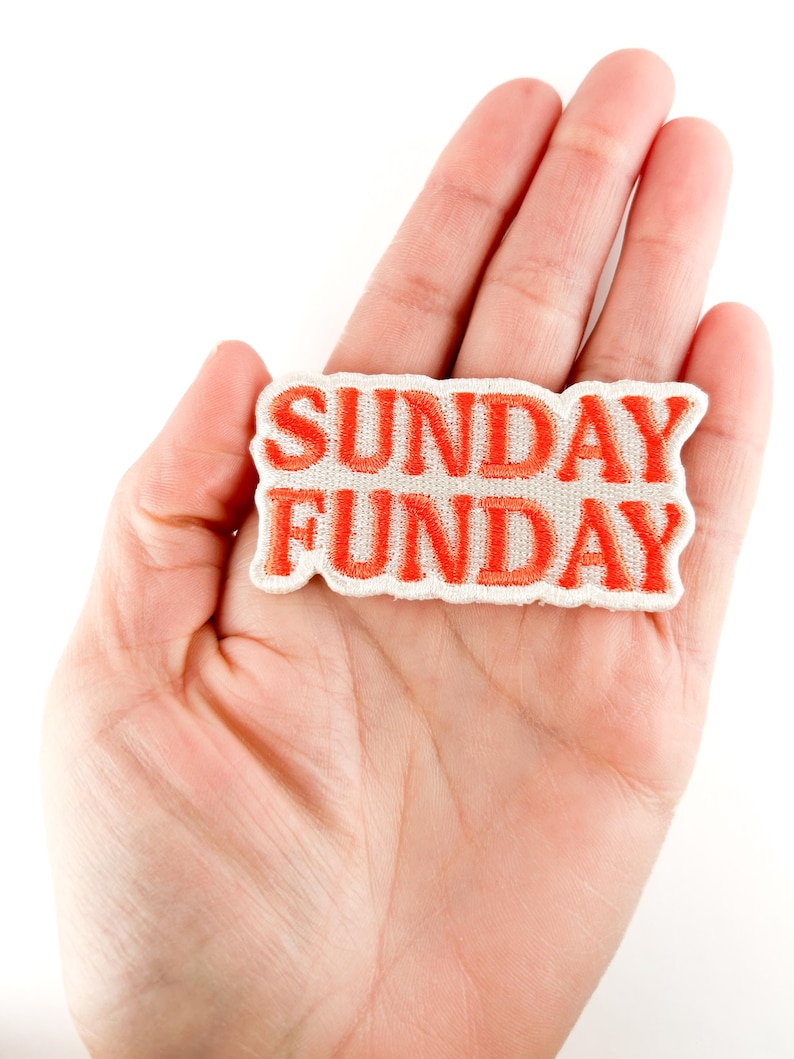 Puede incluir: Un parche rectangular con las palabras "SUNDAY FUNDAY" bordadas en hilo naranja brillante sobre un fondo de tela blanca. El parche se sostiene en la mano de una persona sobre un fondo blanco.
