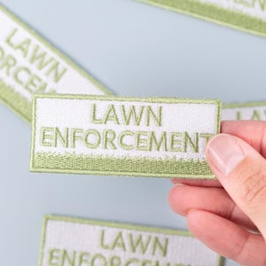 Puede incluir: Parche rectangular bordado en blanco y verde con las palabras "LAWN ENFORCEMENT". El parche está sostenido por una mano. Varios otros parches están dispersos sobre una superficie azul claro.
