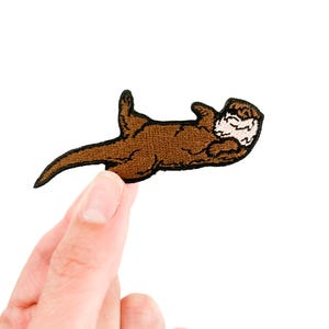 May include: A brown embroidered patch featuring a stylized otter lying on its back. The otter has a white face and is in a relaxed pose. The patch is held against a white background.
