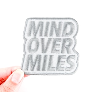 Puede incluir: Un parche bordado gris con las palabras "MIND OVER MILES" en un diseño apilado. El parche tiene un borde blanco y se presenta sobre un fondo blanco.