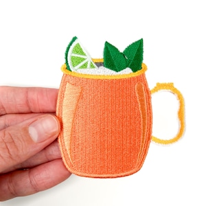 Puede incluir: Un parche bordado de un vaso de cobre con una rodaja de lima y hojas de menta. El vaso es naranja con un borde y un asa amarillos.