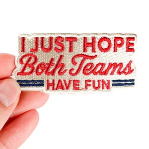 K&ouml;nnte beinhalten: Gestickter Aufn&auml;her mit dem Text "I JUST HOPE Both Teams HAVE FUN" in roter Schrift auf beigem Hintergrund. Zwei blaue Streifen befinden sich am unteren Rand. Der Aufn&auml;her ist rechteckig mit dekorativem Rand.
