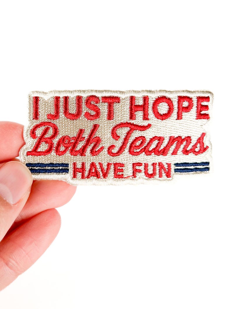 K&ouml;nnte beinhalten: Gestickter Aufn&auml;her mit dem Text "I JUST HOPE Both Teams HAVE FUN" in roter Schrift auf beigem Hintergrund. Zwei blaue Streifen befinden sich am unteren Rand. Der Aufn&auml;her ist rechteckig mit dekorativem Rand.