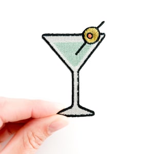 May include: Embroidered patch featuring a martini glass with a green liquid and an olive on a toothpick. The patch has a black outline and a white background. The olive is yellow with a brown center.