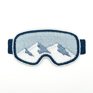 May include: An embroidered patch shaped like ski goggles with a dark blue border. The lens area features a light blue background with white and dark blue mountain peaks, creating a reflective effect. The patch has small rectangular side tabs.