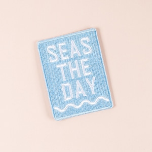 Op de afbeelding: Een rechthoekige, lichtblauwe patch met de tekst "SEAS THE DAY" in het wit geborduurd. Onderaan een witte golflijn. De patch ligt op een lichtperzikkleurige achtergrond.