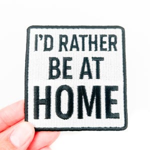 May include: Square white patch with black embroidered text that reads "I'D RATHER BE AT HOME". The patch has a black border and is held by a hand against a white background.