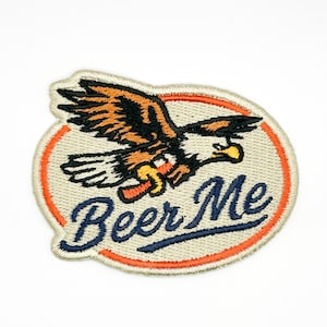 May include: An oval-shaped embroidered patch with a cream-colored background and an orange border. The patch features a flying eagle in brown, black, and white, clutching a small orange object. The text Beer Me is stitched in dark blue.