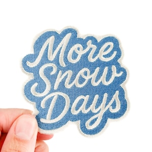 Peut inclure: Écusson brodé bleu en forme de nuage avec l'inscription blanche "More Snow Days". Le patch est tenu par une main sur fond blanc.