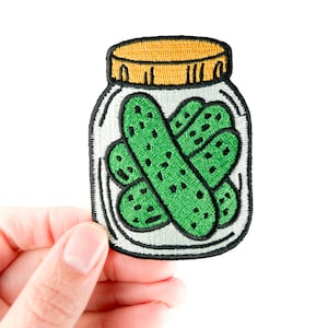 Könnte beinhalten: Gestickter Aufnäher mit einem Glas Gurken. Der Aufnäher zeigt ein klares Glas mit gelbem Deckel und schwarzer Umrandung. Im Inneren des Glases befinden sich mehrere grüne Gurken mit schwarzen Details. Der Aufnäher wird von einer Hand vor weißem Hintergrund gehalten.