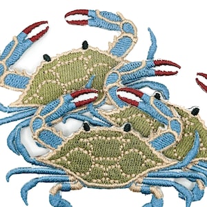 May include: A collection of embroidered crab patches featuring olive green shells with tan outlines and bright blue legs and claws. The claws have distinct red tips and white accents. Each crab has two small black eyes on the top of the shell.