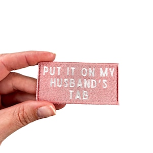 Puede incluir: Un parche de tela rosa con bordado blanco que dice "PUT IT ON MY HUSBAND'S TAB".