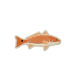 May include: An embroidered patch in the shape of a fish with orange scales, a white underbelly, and a distinct black spot near the tail. The design features detailed stitching throughout the body, fins, and head, finished with a light beige border.