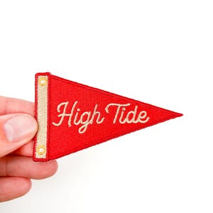 以下が含まれることがあります： 赤いペナント型のパッチで、筆記体で「High Tide」の文字が刺繍されています。 パッチにはクリーム色の縁取りと金色のハトメが付いています。 パッチは白い背景に対して手で持たれています。