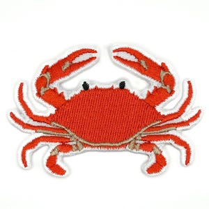 May include: An embroidered iron-on patch shaped like a crab. The crab features a vibrant orange body with white highlights on the claws, small black eyes, and a white border edge. The stitching is dense and textured, creating a detailed crustacean.