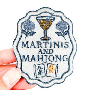 Pode incluir: Patch bordado com fundo branco e borda azul marinho. O patch apresenta um copo de Martini, flores azuis e as palavras "MARTINIs AND MAHJONG". Duas peças de Mahjong na parte inferior.