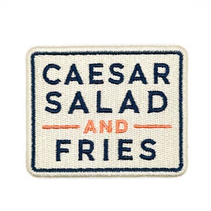 Puede incluir: Un parche bordado rectangular con fondo color crema y borde azul oscuro. El texto CAESAR SALAD está bordado en azul oscuro, seguido de la palabra AND en naranja y FRIES en azul oscuro, con líneas horizontales de color naranja.
