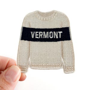 May include: A beige embroidered patch in the shape of a sweater. The patch features a navy blue stripe with the word "VERMONT" in white block letters. The patch is held by a hand against a white background.
