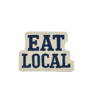 May include: Embroidered patch with the words "EAT LOCAL" in navy blue block letters. The text is on a cream-colored background with a navy blue border. The patch is rectangular with a slightly irregular shape.