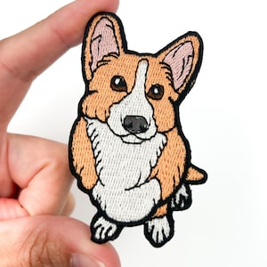 Könnte beinhalten: Gestickter Aufnäher mit einem Cartoon-Corgi-Hund. Der Aufnäher hat eine weiße Brust und ein weißes Gesicht mit hellbraunem und braunem Fell. Der Corgi hat schwarze Augen und eine schwarze Nase. Der Aufnäher wird von einer Hand vor weißem Hintergrund gehalten.