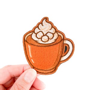 May include: Embroidered patch of a coffee cup with whipped cream. The cup is orange with a brown outline and a white handle. The whipped cream is white with brown accents. The patch is held by a hand against a white background.