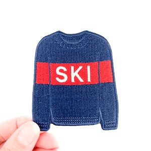 Puede incluir: Parche bordado azul marino con forma de suéter. Presenta una franja roja con la palabra "SKI" en blanco. El parche se sostiene sobre un fondo blanco. Un accesorio divertido para los amantes del esquí.