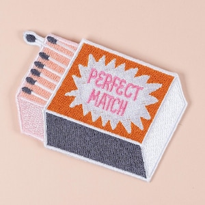 以下が含まれることがあります： 開いたマッチ箱の形をした刺繍パッチ。マッチ箱はオレンジ、グレー、白で、「PERFECT MATCH」の文字がピンク色で書かれています。マッチはピンクと黒です。