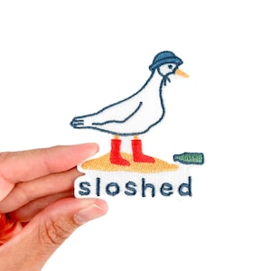 May include: Embroidered patch featuring a white goose wearing a blue hat and red boots, standing on a sandy area with a green bottle. The word "sloshed" is below the goose.