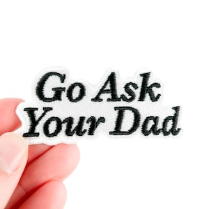 May include: Embroidered patch with the phrase "Go Ask Your Dad" in black script on a white background. The patch is held by a hand against a white backdrop. This patch is suitable for jackets, bags, or other fabric items.