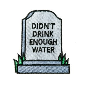 May include: Embroidered patch in the shape of a tombstone with the words "DIDN'T DRINK ENOUGH WATER" in black. The tombstone is light gray with a dark gray border and green grass at the bottom.