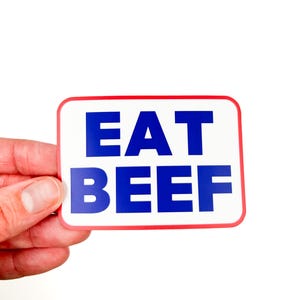 May include: A white sign with a red border and blue text that reads "EAT BEEF".