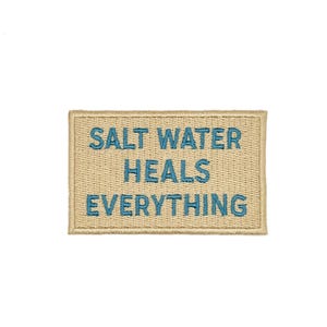 Puede incluir: Un parche rectangular con el texto bordado "SALT WATER HEALS EVERYTHING" en azul sobre un fondo beige. El parche tiene un borde cosido y probablemente está diseñado para ser cosido en ropa o accesorios.