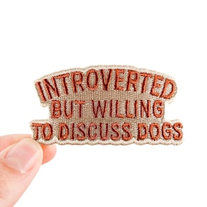 Op de afbeelding: Geborduurde patch met de tekst "INTROVERTED BUT WILLING TO DISCUSS DOGS" in oranje draad op een beige achtergrond. De patch wordt door een hand tegen een witte achtergrond gehouden.