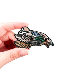 May include: An embroidered patch of a brown and green duck in flight. The duck has a light blue wing and orange feet.