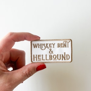 May include: A rectangular white patch with gold embroidered text that reads "WHISKEY BENT & HELLBOUND". The patch has a gold border and is held by a hand with red nail polish.