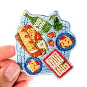 May include: Embroidered patch depicting a picnic scene. The patch shows a green bottle, bread, strawberries, and small plates of food, set on a blue checkered background. The patch is held by a hand.