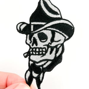 May include: Black and white embroidered patch featuring a skull wearing a cowboy hat and smoking a cigar. The patch has a detailed design with a classic tattoo style. The skull is white with black outlines and shading.