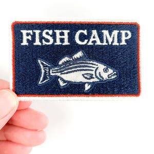 May include: Embroidered patch featuring the text "FISH CAMP" in white above a white fish illustration. The patch has a navy blue background with a red border. The patch is rectangular in shape.