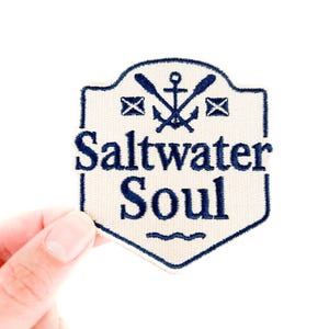 May include: A cream-colored patch with a navy blue border and text that reads "Saltwater Soul." The patch features an anchor, oars, and envelope icons. The patch is held by a hand against a white background.