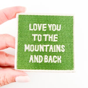 Puede incluir: Parche de tela verde con texto bordado en blanco que dice "Love you to the mountains and back".
