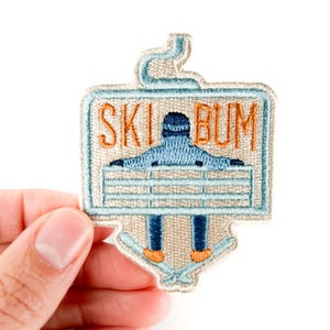 May include: Embroidered patch with the words "SKI BUM" in orange lettering. The patch features a skier in a blue outfit with orange boots and skis. The patch is in a light blue and beige color scheme.