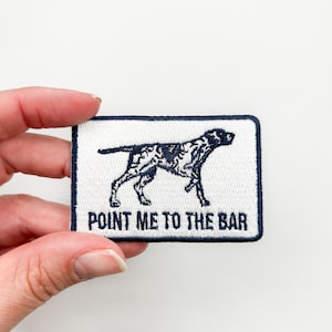 May include: White embroidered patch with a black and white dog pointing to the right. The text "POINT ME TO THE BAR" is below the dog.