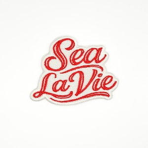 May include: Embroidered patch with the words "Sea La Vie" in red script lettering. The text is on a white background with a red border. The patch has a nautical or beach theme.