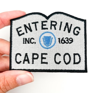Könnte beinhalten: Gestickter Aufnäher mit hellgrauem Hintergrund und schwarzem Rand. Der Aufnäher trägt den Text "ENTERING INC. CAPE COD 1639" in schwarzer Schrift. Ein blaues Emblem befindet sich in der Mitte. Der Aufnäher wird von einer Hand gehalten.